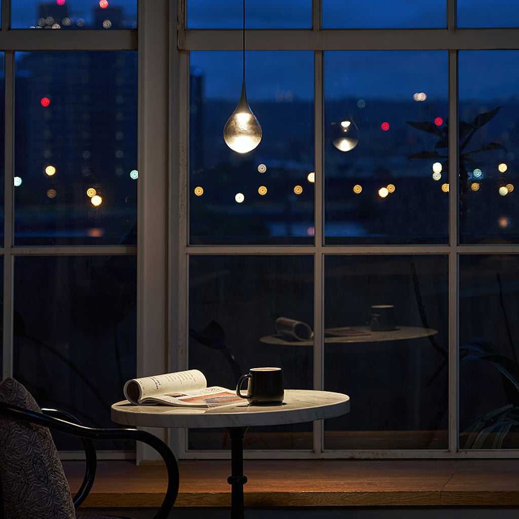 A single Clérachic pendant with a clear teardrop shade glows in a window at night, above a small table with an open book.