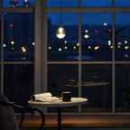 A single Clérachic pendant with a clear teardrop shade glows in a window at night, above a small table with an open book.