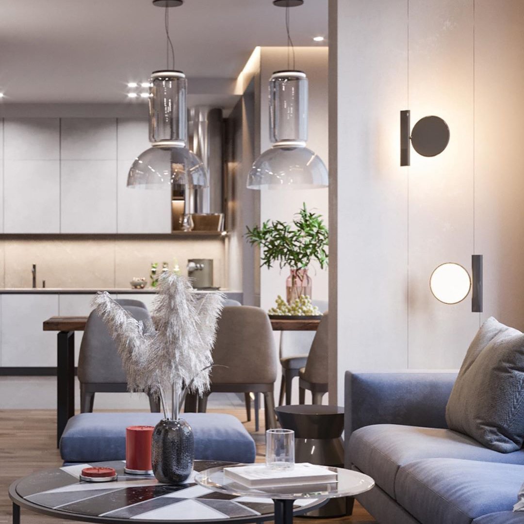 Two Clérachic glass pendants over a kitchen and living area, with a wall light, a blue sofa and a vase of pampas grass.