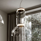 Looking up at this Clérachic pendant's glass cylinder, lit ring and dome, beside a window with leaves and curtains.