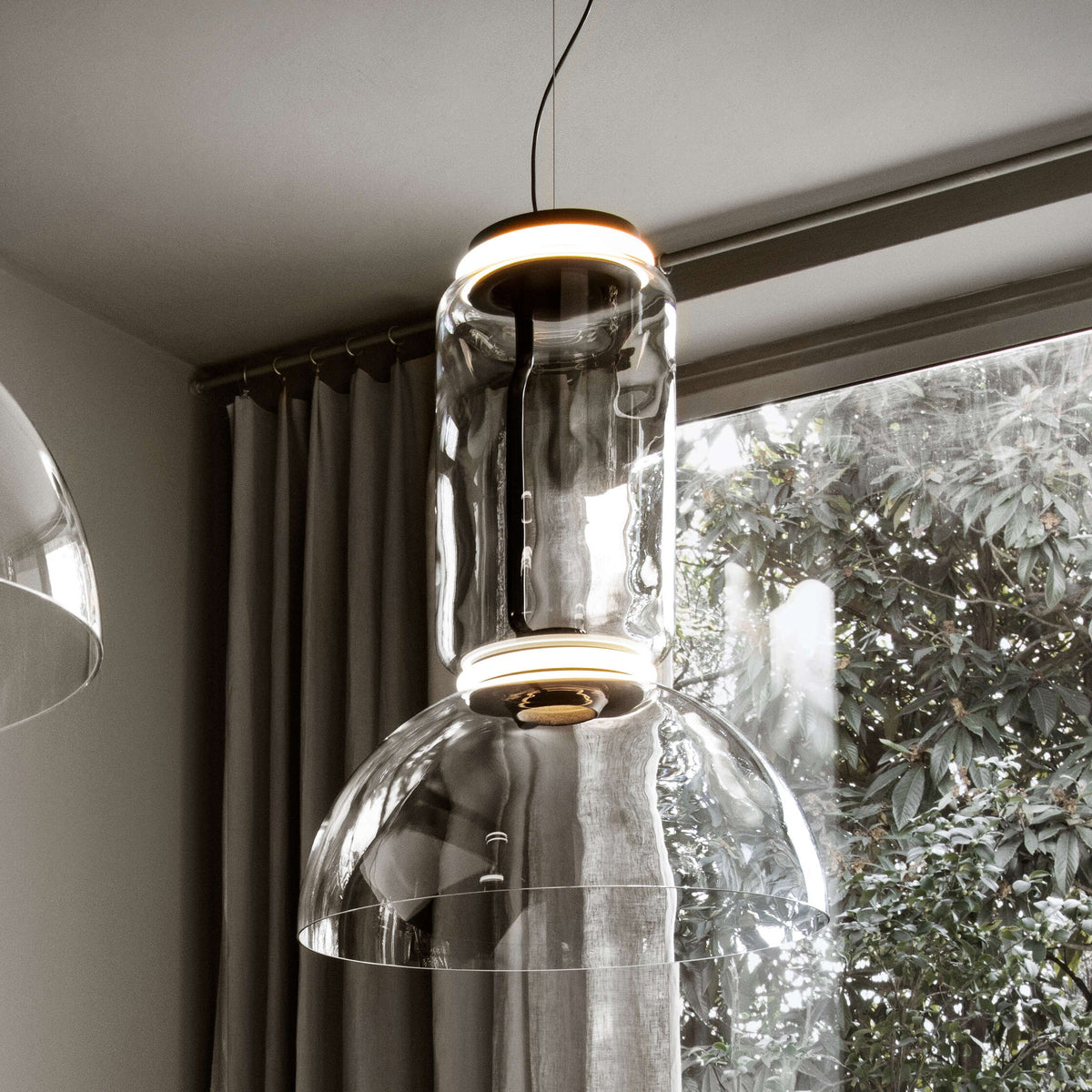 Looking up at this Clérachic pendant's glass cylinder, lit ring and dome, beside a window with leaves and curtains.