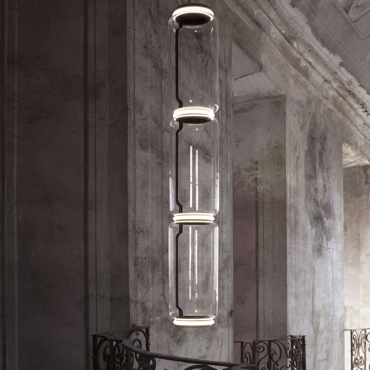 Tall stack of glass cylinders with lit rings, a Clérachic pendant seen in front of a stone column and a balcony rail.