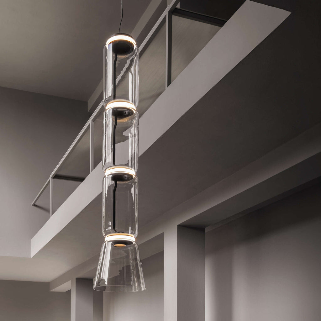 Four stacked glass cylinders and a cone, this Clérachic pendant hanging past a mezzanine in a grey, modern room.