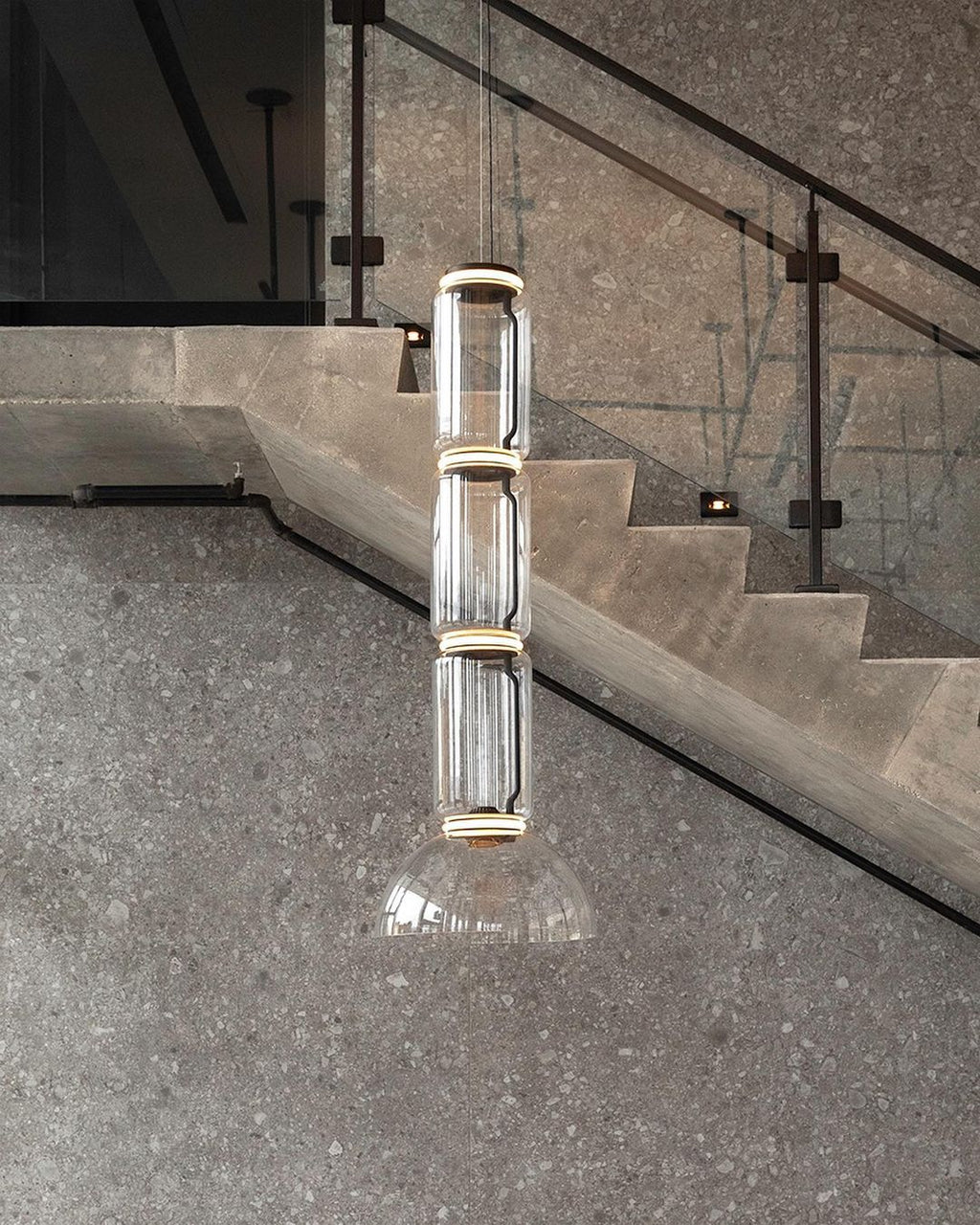 Stacked glass cylinders over a cone, this Clérachic pendant lit beside a stone staircase with a glass balustrade.