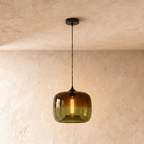Clérachic glass pendant in amber fading to olive green, lit by a warm filament bulb, hung on a black cord from a black canopy.