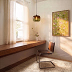 Clérachic amber and green glass pendant glowing above a walnut floating desk, with a leather cantilever chair and sheer curtains.