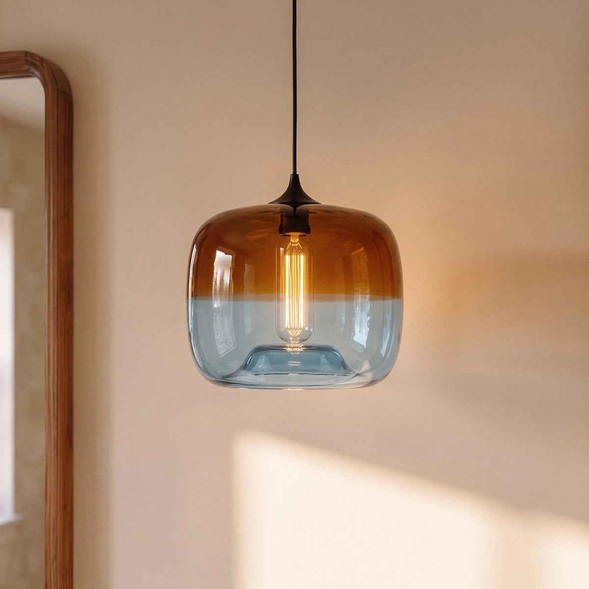 Close-up of the Clérachic amber-to-blue glass pendant, its filament bulb glowing warm against a sunlit cream wall.