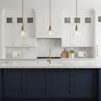 Three Clérachic clear glass pendants with brass fittings hang above a white kitchen's marble counter and a navy island.