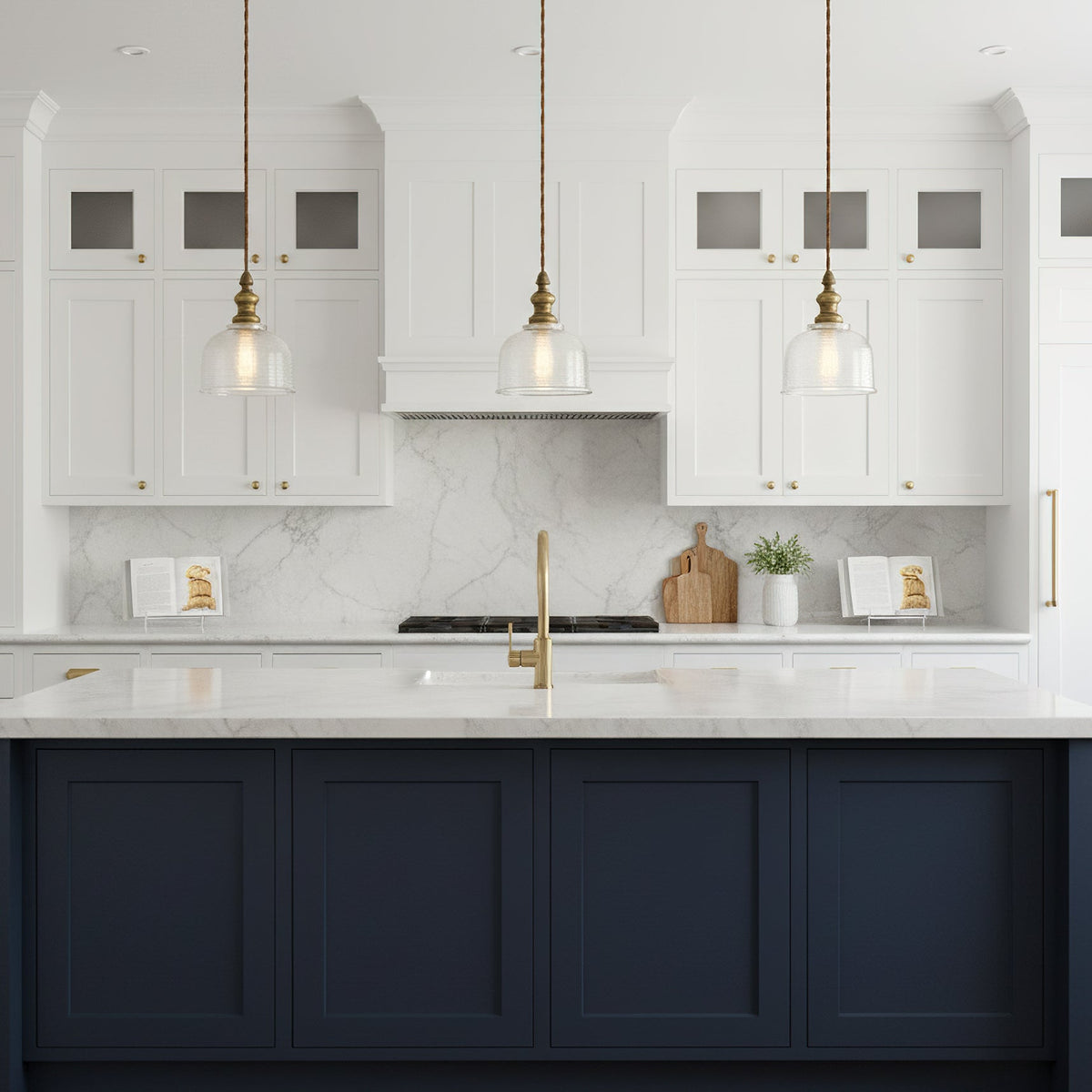 Three Clérachic clear glass pendants with brass fittings hang above a white kitchen's marble counter and a navy island.
