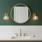 Two glass-shade brass wall lights from Clérachic flank a round brass mirror on a green wall above a sink with brass taps.
