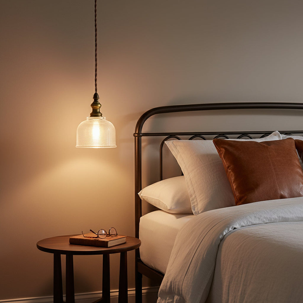 A Clérachic ribbed glass pendant glows beside a bed with a black metal headboard, leather cushions and a round bedside table.