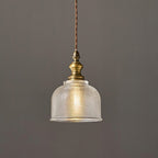 Close view of a Clérachic pendant's ribbed glass dome and turned antique brass fitting, with a twisted cord, against a grey wall.