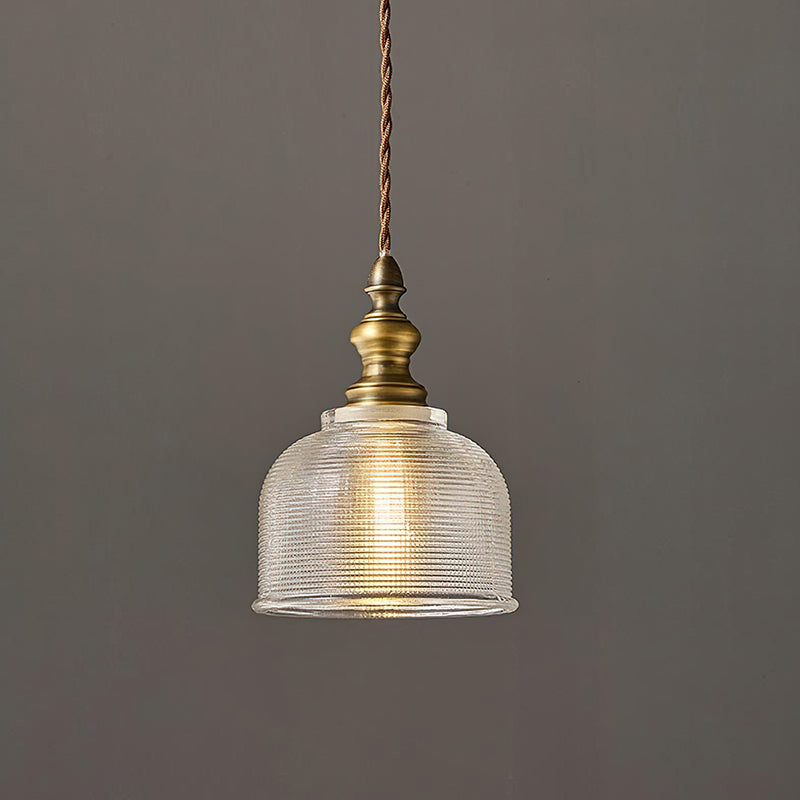 Close view of a Clérachic pendant's ribbed glass dome and turned antique brass fitting, with a twisted cord, against a grey wall.