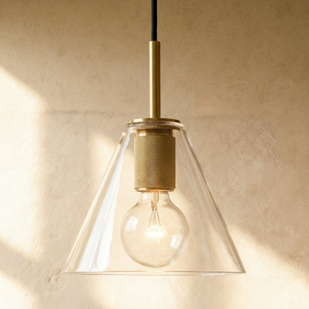 Clérachic brass pendant light with a clear cone-shaped glass shade over an exposed bulb against a plaster wall.