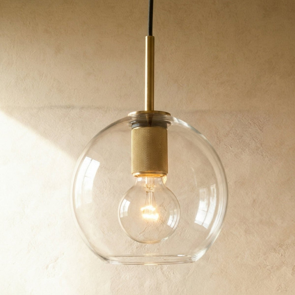 Clérachic brass pendant light with a clear spherical glass shade over an exposed bulb against a plaster wall.