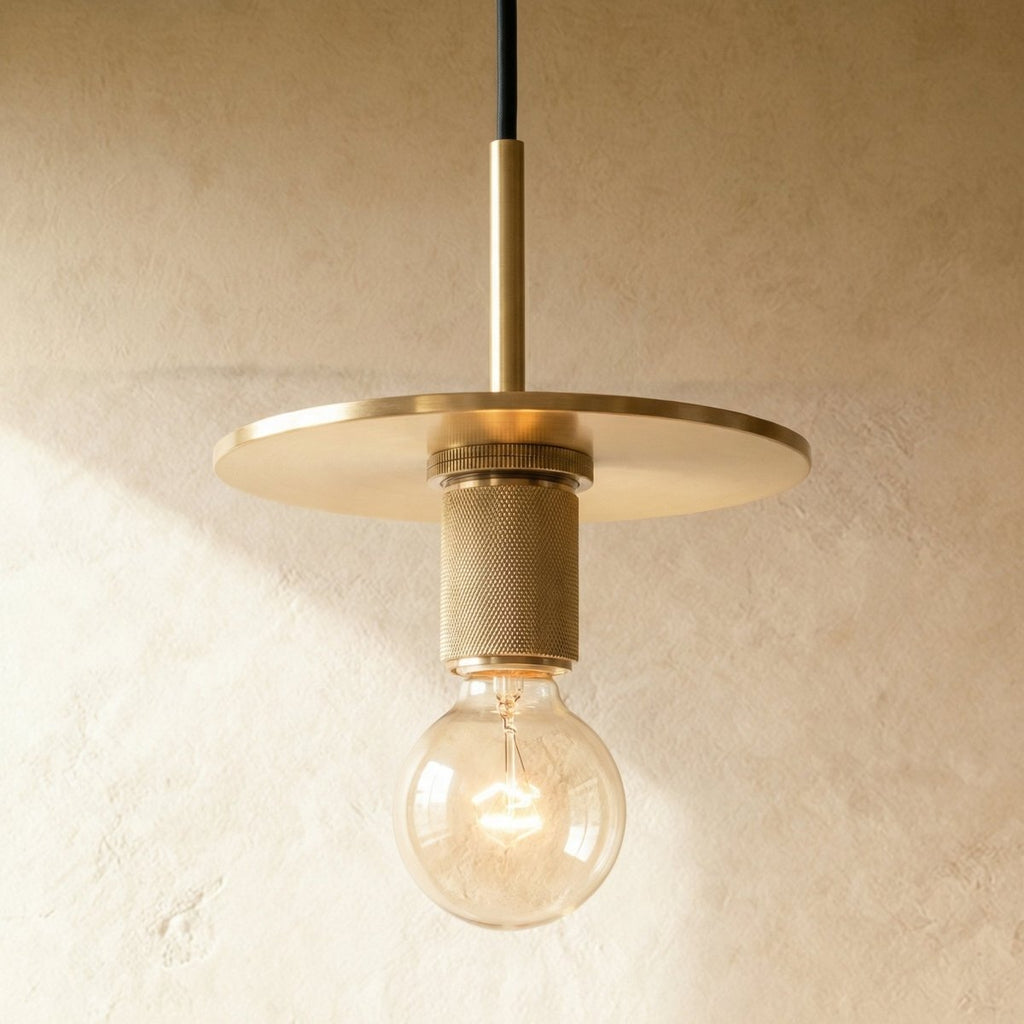 Clérachic brass pendant light with a flat brass disc shade over an exposed bulb against a plaster wall.