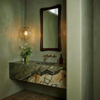 Clérachic brass pendant light with a clear globe shade glowing warm above a green-marble bathroom vanity.