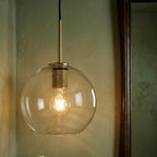Clérachic brass pendant light with a clear globe shade glowing beside a dark wood-framed mirror in a green bathroom.