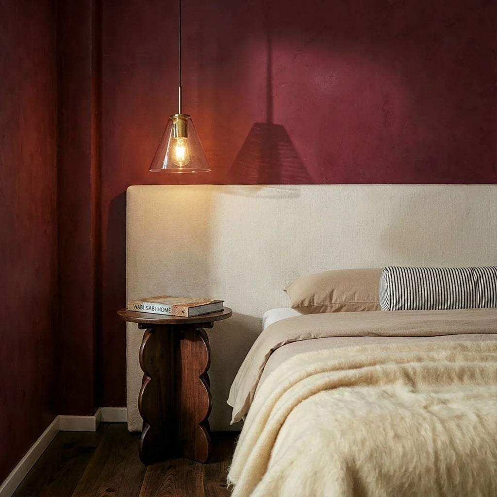Clérachic brass pendant light with a clear cone shade glowing above a walnut nightstand in a maroon bedroom.