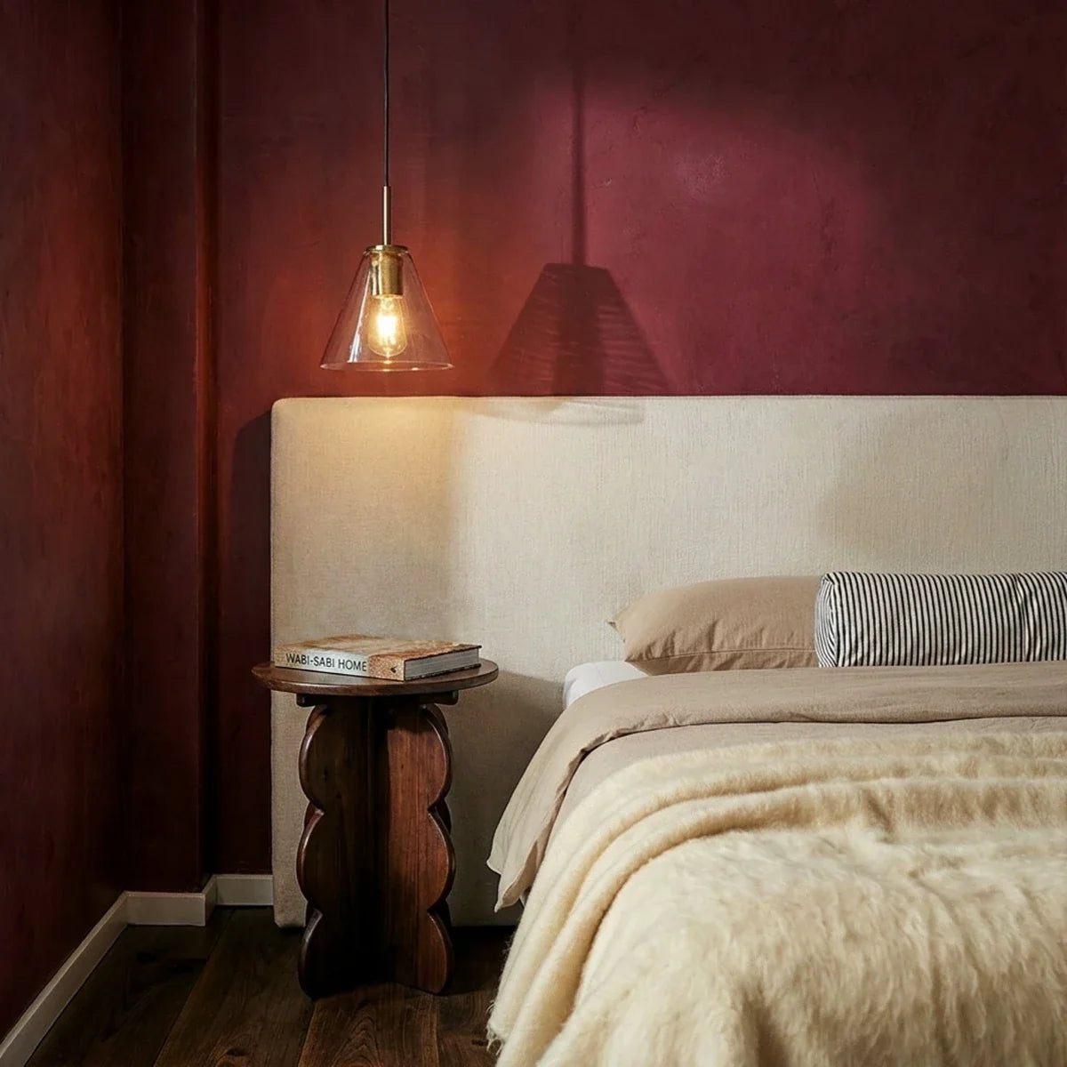 Clérachic brass pendant light with a clear cone shade glowing above a walnut nightstand in a maroon bedroom.