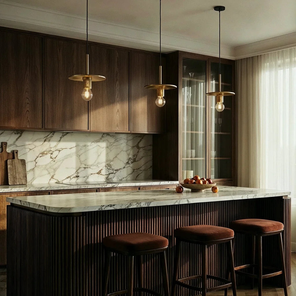Clérachic brass disc-shade pendant lights glowing in a row above a marble kitchen island with bar stools.