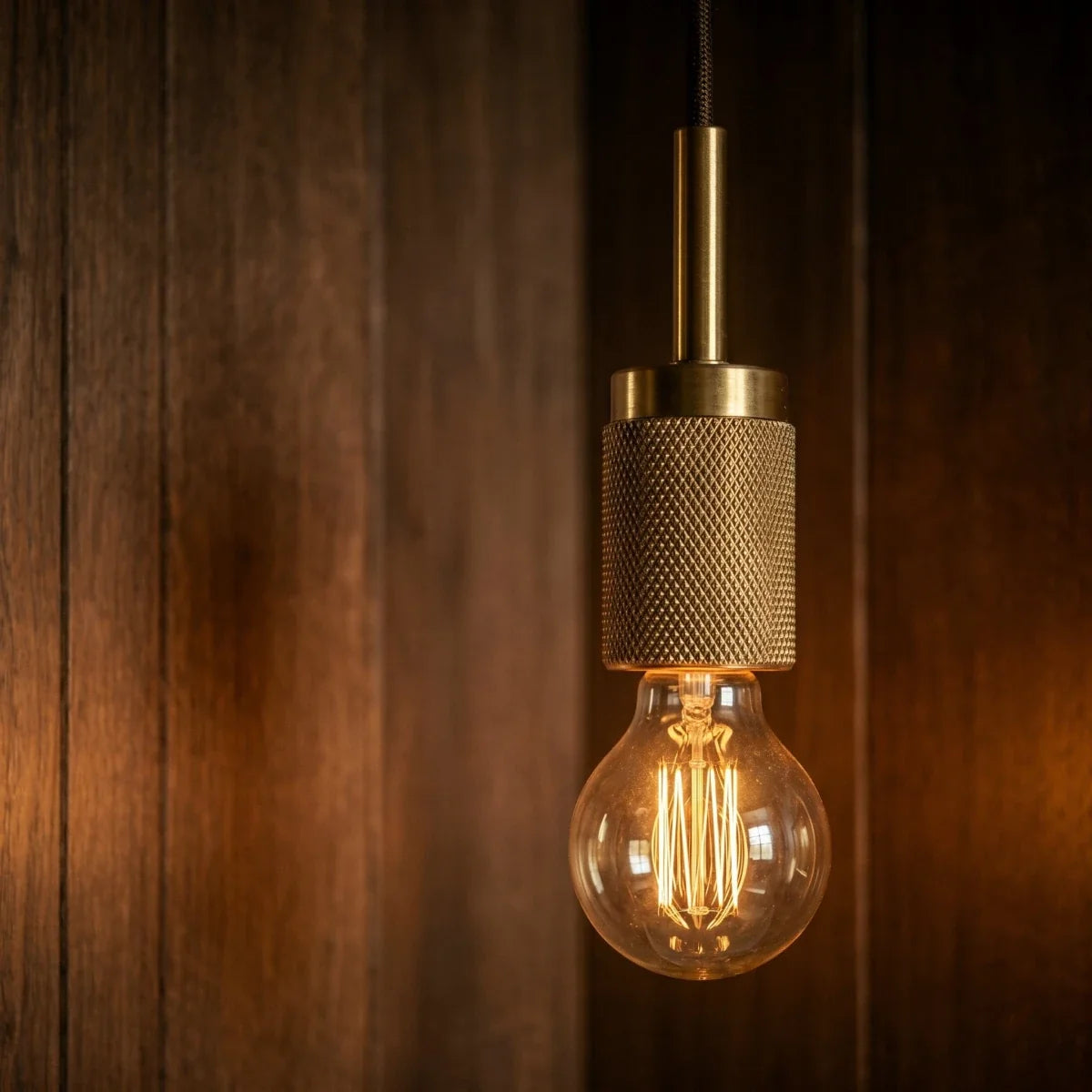 Clérachic brass pendant light in close detail, its knurled socket and exposed bulb glowing against wood paneling.