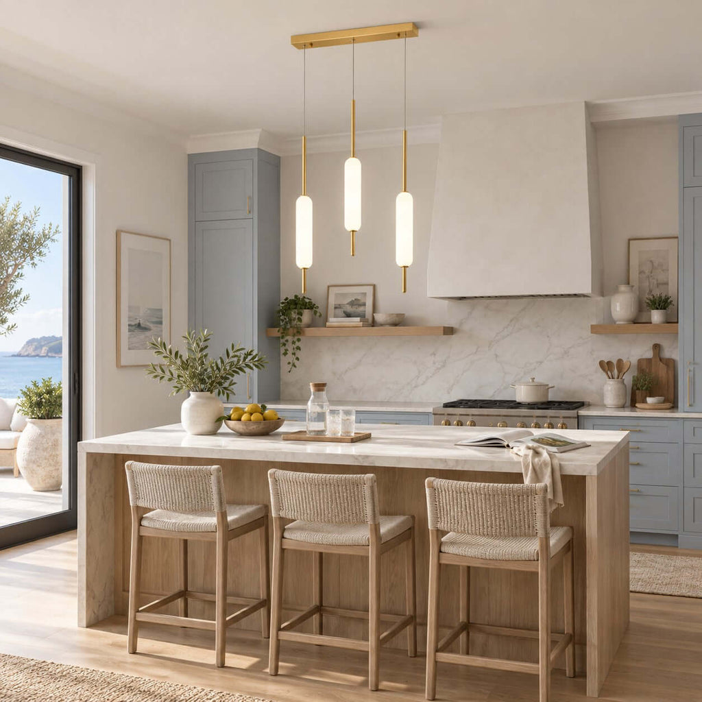 Three Clérachic frosted glass pendants on a brass bar hang over a pale wood kitchen island with woven stools and blue-grey cabinets.