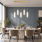 Five Clérachic frosted glass pendants at staggered heights hang from a brass bar over a marble dining table with taupe chairs.