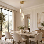 A clustered Clérachic glass pendant on a round brass canopy hangs by tall windows, above a marble dining table with cream chairs.