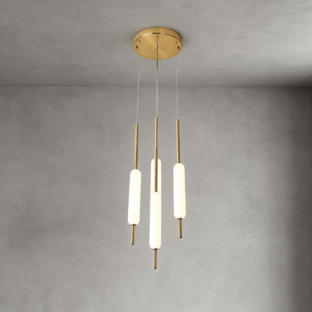 Three lit Clérachic frosted glass pendants on brass rods hang from a round brass canopy against a grey plaster wall.