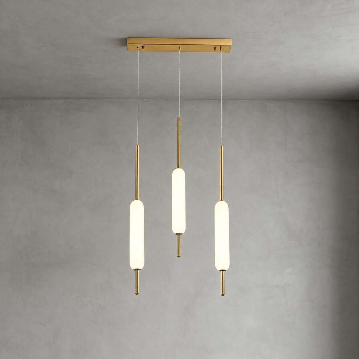 Three Clérachic frosted glass pendants at different heights hang from a slim brass bar against a grey plaster wall.
