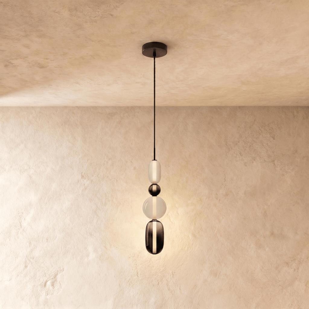 Clérachic tall pendant in frosted glass, a dark bead, an opal globe and a smoked capsule, lit on a plaster wall.