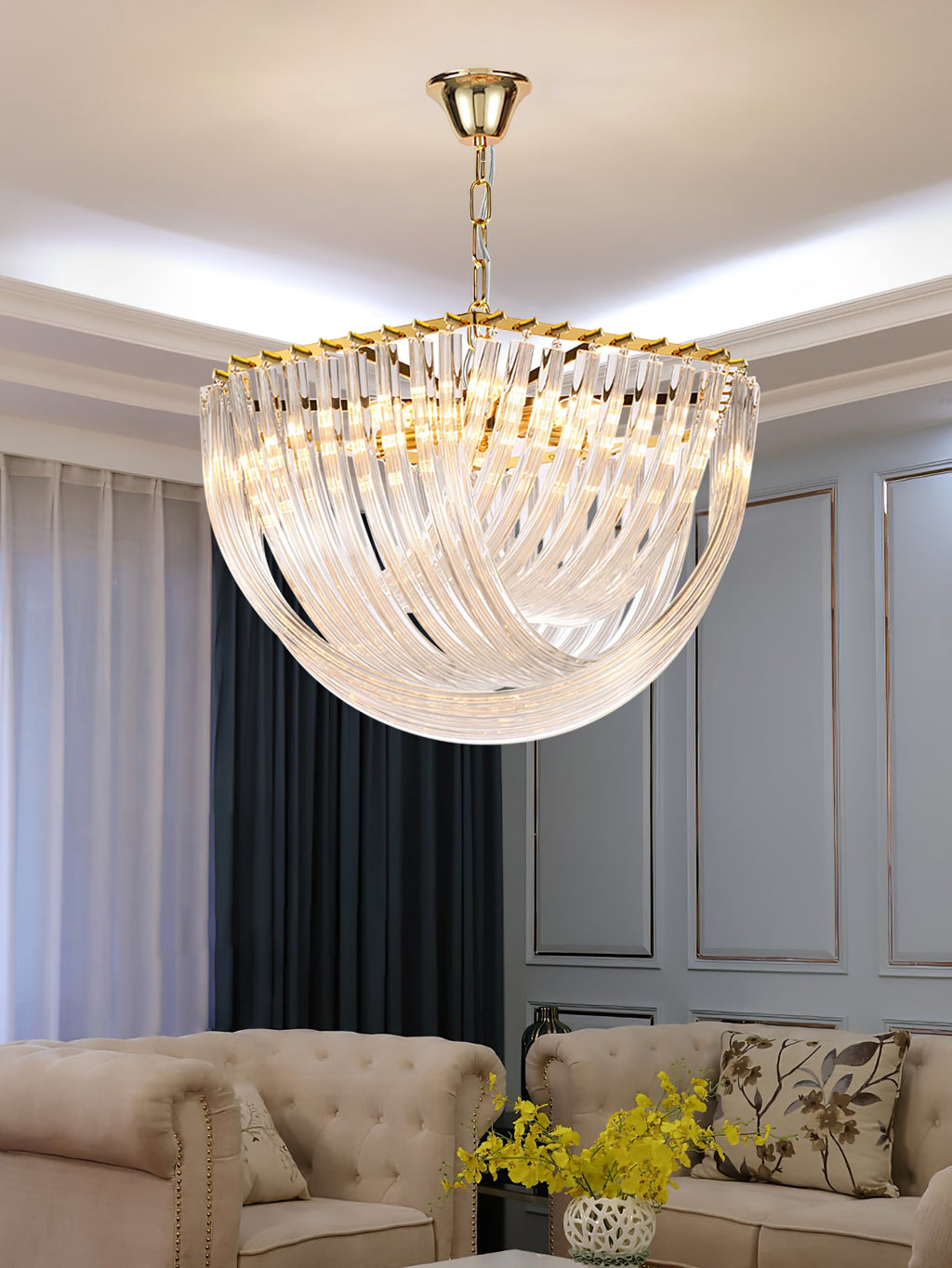 Clérachic chandelier on a gold chain above a cream tufted sofa, with grey curtains and yellow flowers in the foreground.