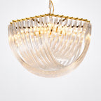 Close angled view of the curved clear glass rods and gold edge of this Clérachic chandelier, lit warmly on a pale backdrop.