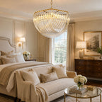 Clérachic chandelier lit above a bedroom with a cream upholstered bed, a gold-framed painting and a lamp on a wooden chest.