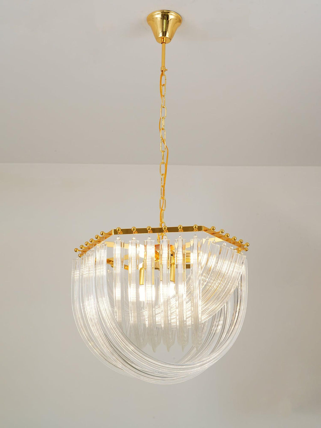 Lit Clérachic chandelier on a gold chain with curved clear glass rods and a hexagonal gold frame, against a pale wall.