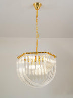 Lit Clérachic chandelier on a gold chain with curved clear glass rods and a hexagonal gold frame, against a pale wall.