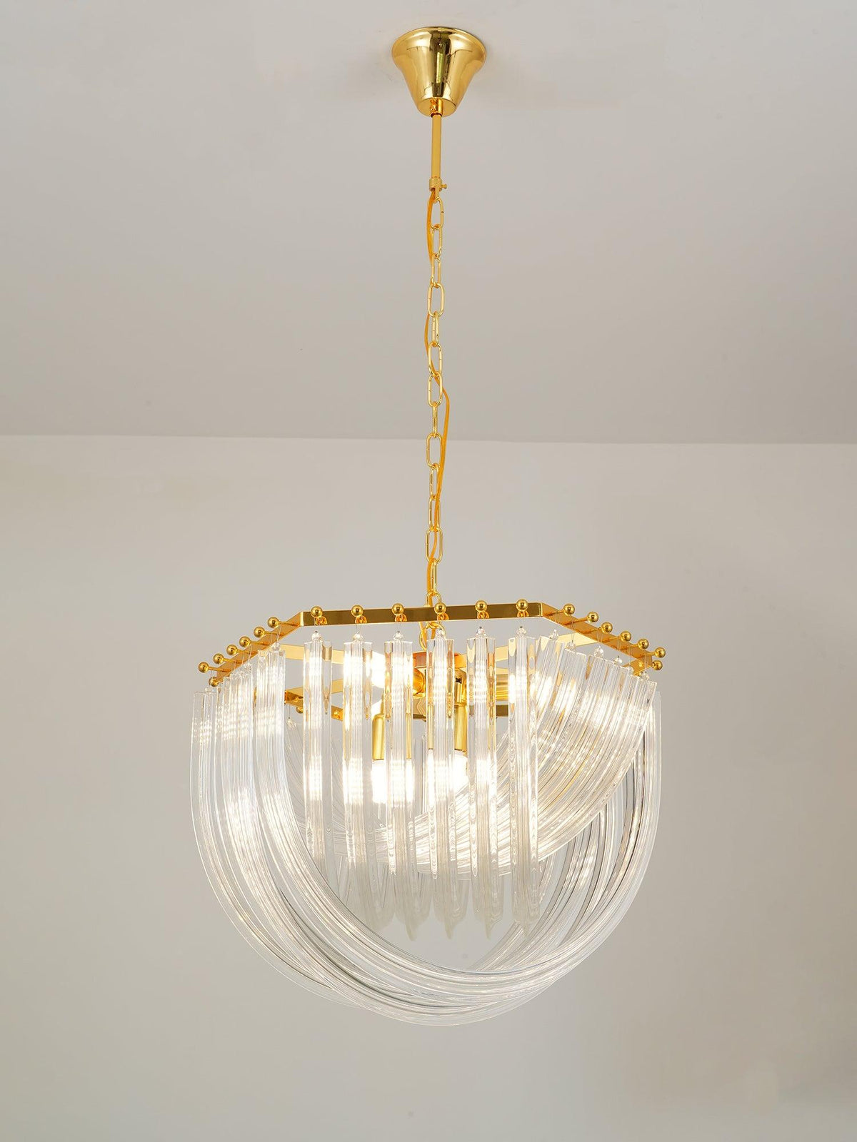 Lit Clérachic chandelier on a gold chain with curved clear glass rods and a hexagonal gold frame, against a pale wall.