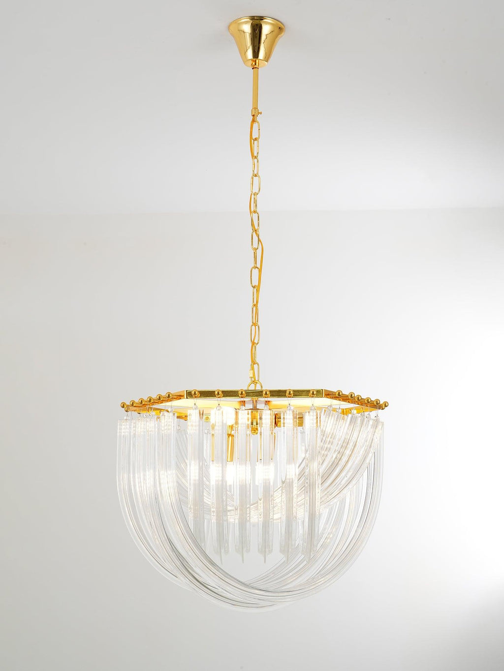 Clérachic chandelier hung from a long gold chain, its looped clear glass rods and gold frame seen in a pale, plain room.