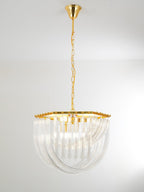 Clérachic chandelier hung from a long gold chain, its looped clear glass rods and gold frame seen in a pale, plain room.