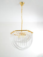Unlit Clérachic chandelier with a hexagonal gold top and clear looped glass rods, hanging from a gold chain on a plain wall.