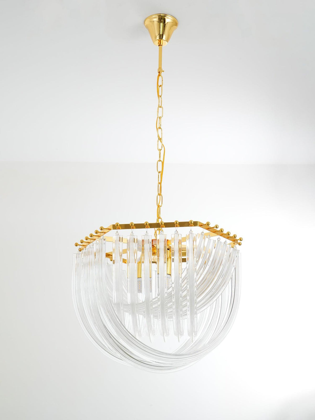 Unlit Clérachic chandelier with a hexagonal gold top and clear looped glass rods, hanging from a gold chain on a plain wall.
