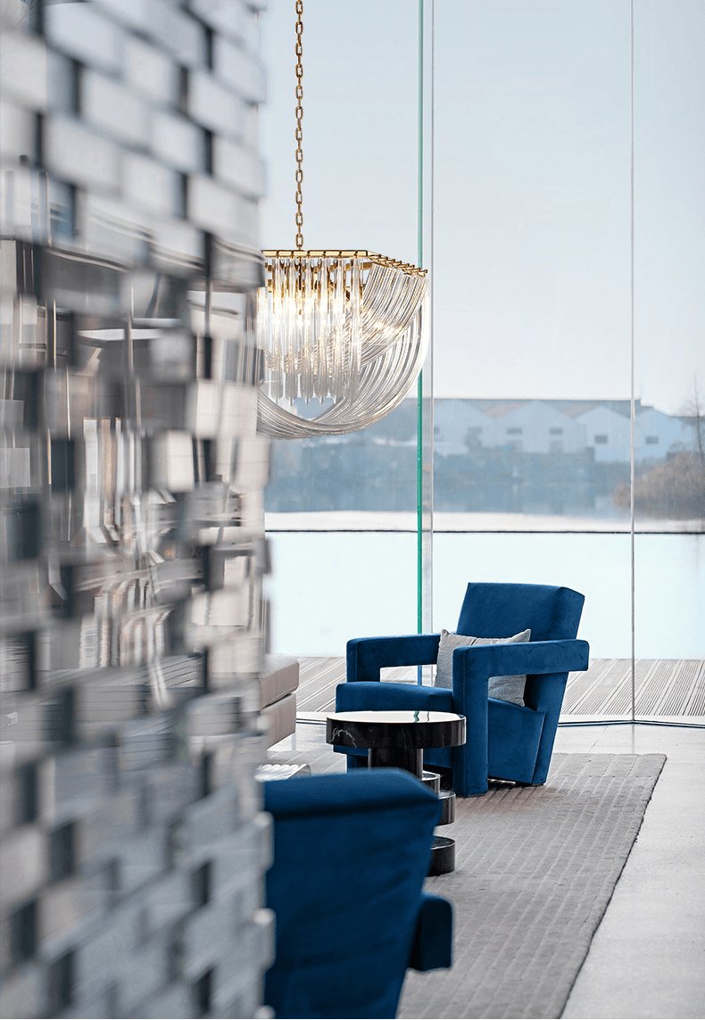 Clérachic chandelier hung beside a floor-to-ceiling window with a lake view, with blue armchairs and a textured wall in front.