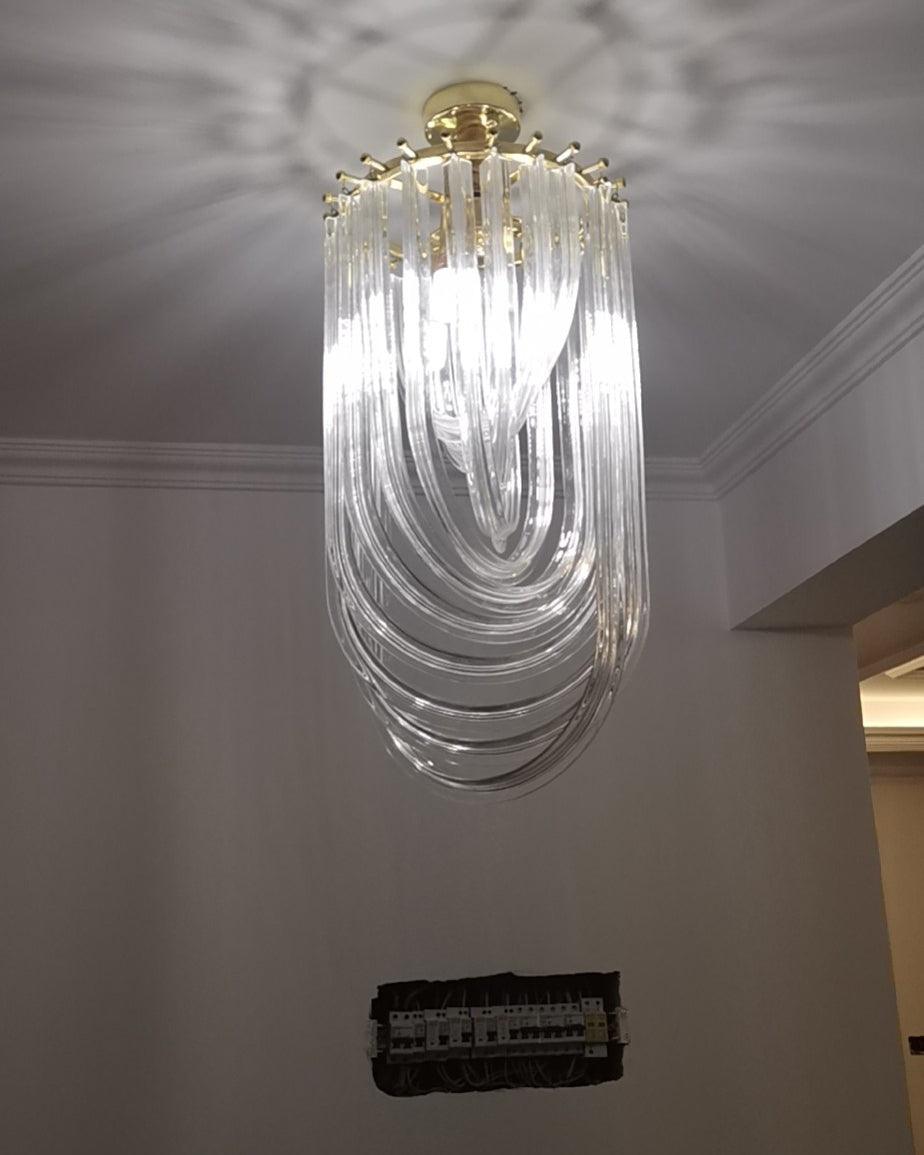 Long, narrow Clérachic chandelier of looped clear glass hung from a gold canopy in a room with a grey wall and cornice.