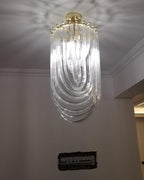 Long, narrow Clérachic chandelier of looped clear glass hung from a gold canopy in a room with a grey wall and cornice.