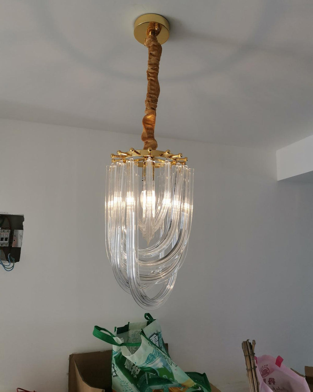 Narrow looped-glass Clérachic chandelier hung from a gold canopy on a twisted fabric cord, with a green bag on the floor below.