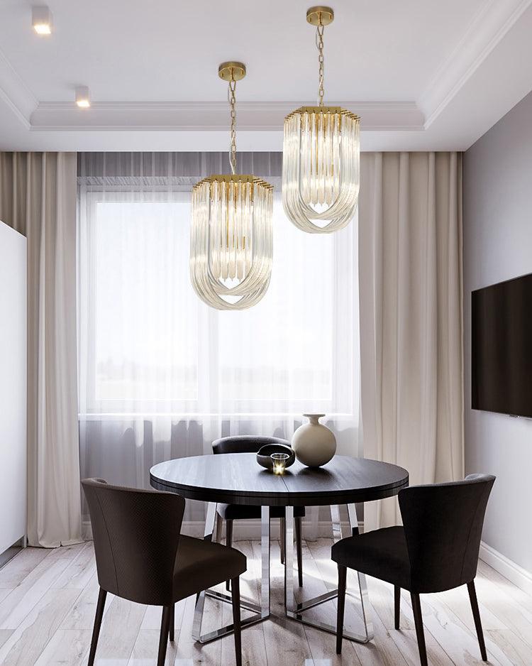 Two narrow looped-glass Clérachic chandeliers hung at different heights over a round dark dining table with brown chairs.