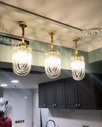 Three narrow looped-glass Clérachic pendants lit in a row above a kitchen with dark cabinets and cornice moulding.