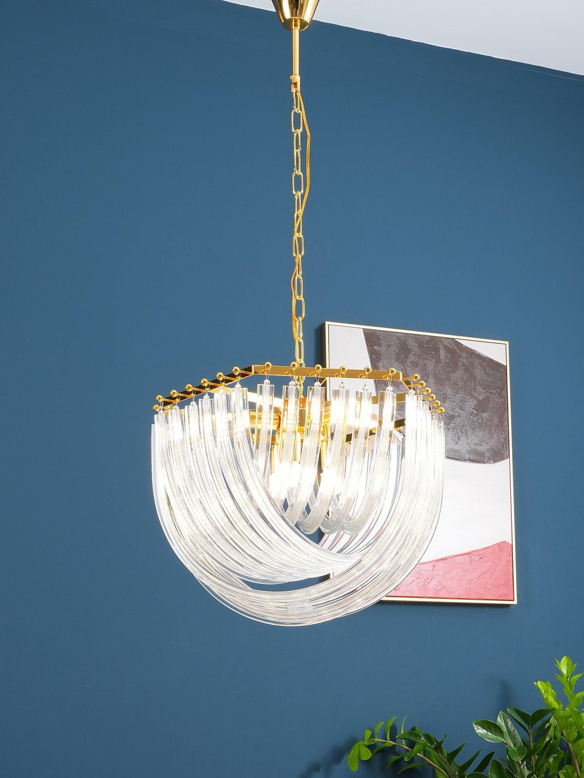 Clérachic chandelier on a gold chain against a blue wall, its looped clear glass rods lit beside an abstract picture.