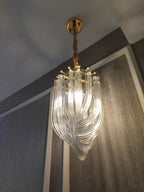 Narrow looped-glass Clérachic chandelier lit from below, hung from a gold canopy in front of a grey textured wall.